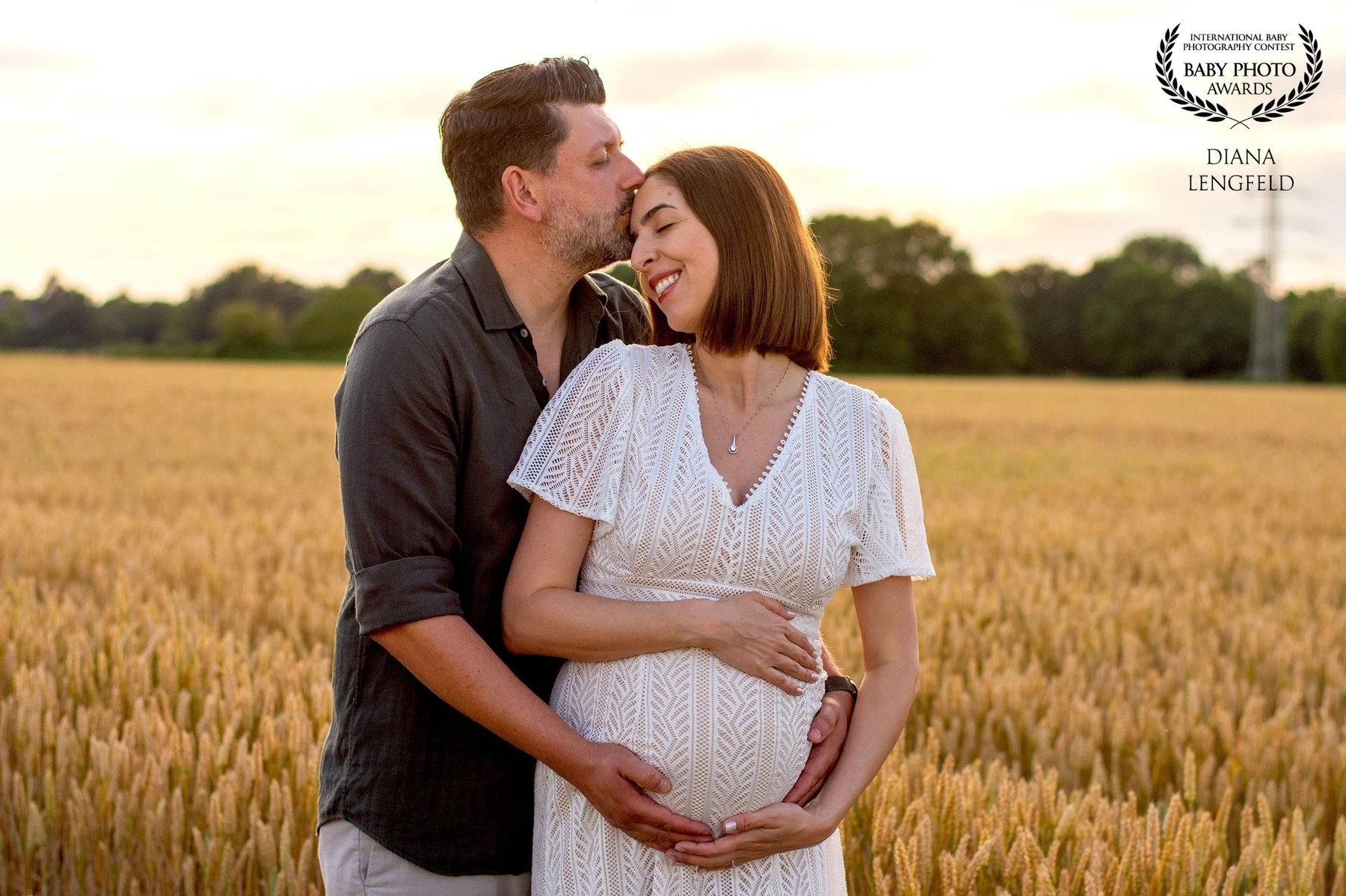 Paar im Kornfeld, er küsst ihre Stirn, beide halten den Babybauch; mit Baby Photo Awards Signet