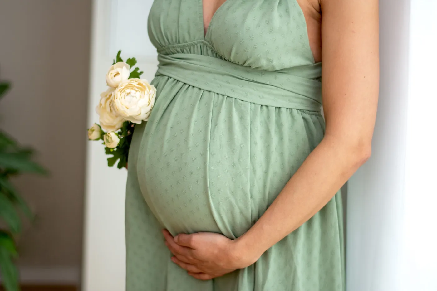 Babybauch in einem salbeigrünen Kleid, daneben cremefarbene Blüten